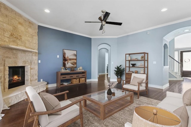 living room featuring baseboards, ornamental molding, a stone fireplace, wood finished floors, and arched walkways