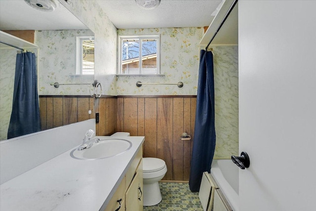 full bathroom with a wainscoted wall, toilet, vanity, a textured ceiling, and wallpapered walls