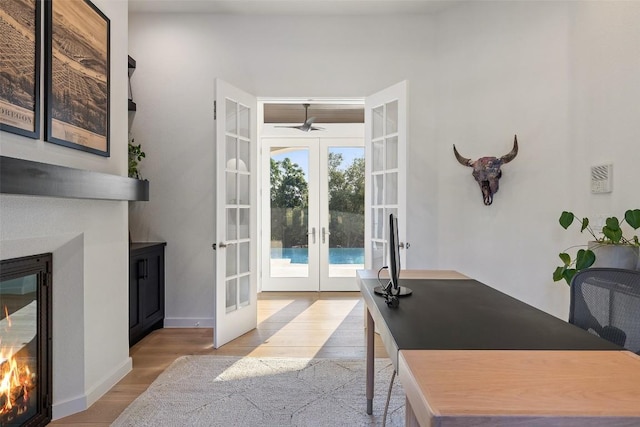 office featuring baseboards, a ceiling fan, a glass covered fireplace, light wood-style flooring, and french doors