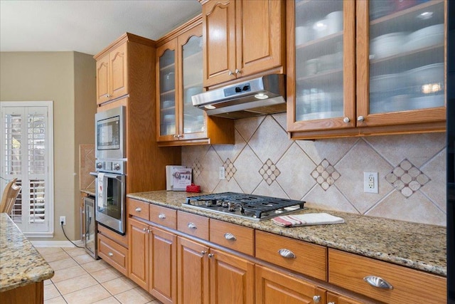 kitchen with stainless steel appliances, light stone counters, brown cabinetry, and under cabinet range hood