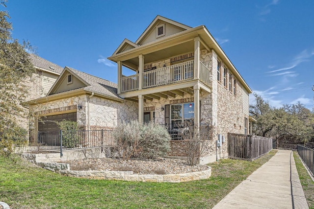 view of front facade featuring stone siding, fence, a front yard, an attached garage, and a balcony