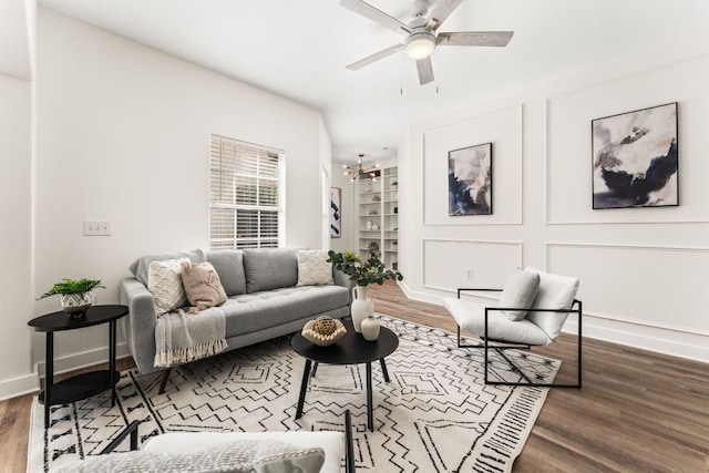 living room featuring built in shelves, a decorative wall, wood finished floors, baseboards, and ceiling fan with notable chandelier