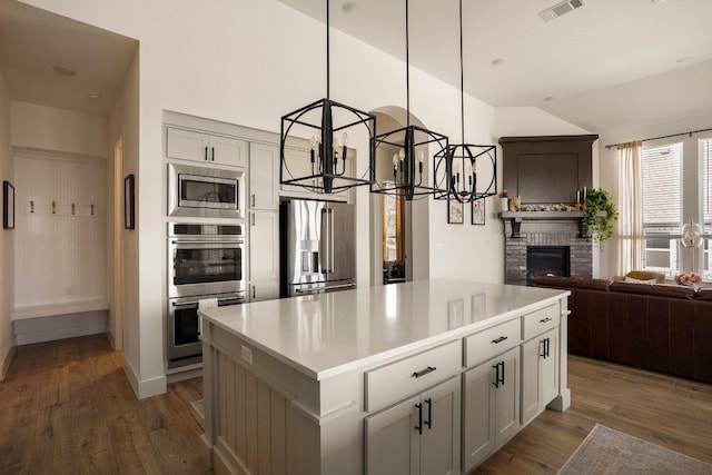 kitchen featuring open floor plan, appliances with stainless steel finishes, a brick fireplace, a center island, and dark wood-style floors