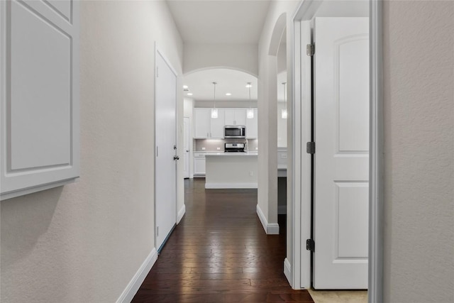 hallway featuring arched walkways, a textured wall, dark wood-style flooring, and baseboards