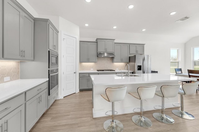 kitchen with a center island with sink, appliances with stainless steel finishes, gray cabinetry, under cabinet range hood, and a sink