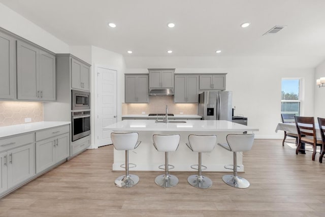 kitchen with under cabinet range hood, a sink, appliances with stainless steel finishes, gray cabinets, and a center island with sink