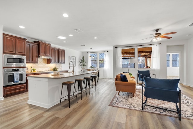 kitchen with a sink, appliances with stainless steel finishes, light wood-type flooring, backsplash, and an island with sink