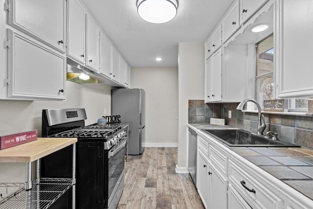 kitchen with tile countertops, under cabinet range hood, stainless steel appliances, a sink, and white cabinetry