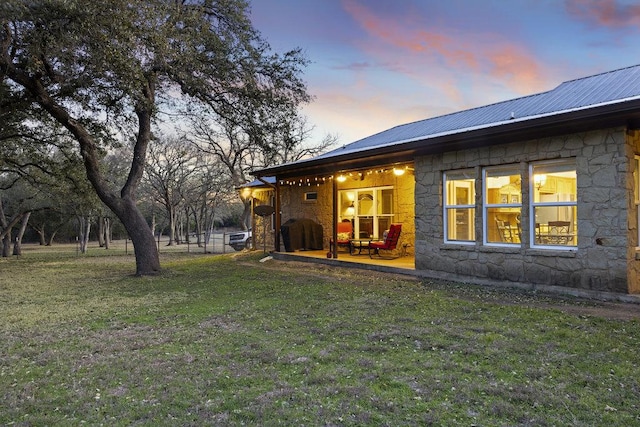 back of house at dusk with a yard, stone siding, and metal roof