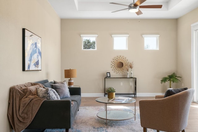 living area with ceiling fan, a tray ceiling, wood finished floors, and baseboards