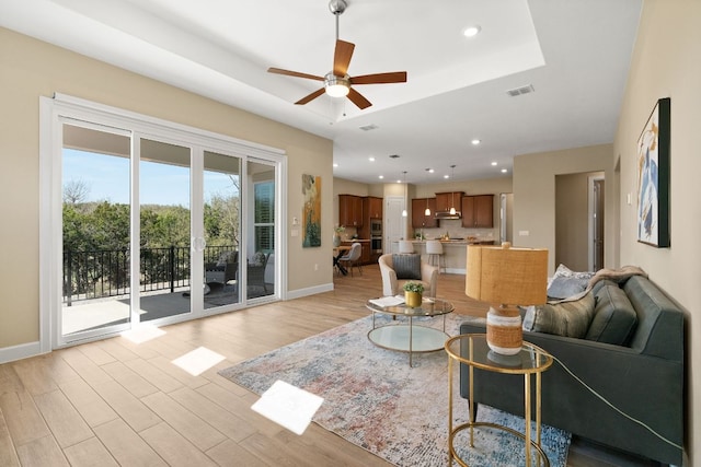 living room with a tray ceiling, light wood finished floors, visible vents, ceiling fan, and baseboards