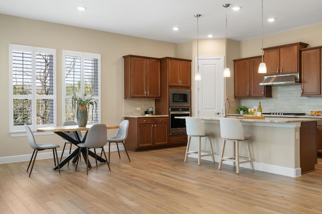 kitchen with brown cabinets, a center island with sink, stainless steel appliances, a sink, and under cabinet range hood