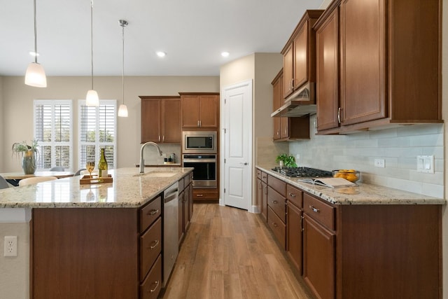 kitchen with brown cabinetry, light wood-style floors, appliances with stainless steel finishes, under cabinet range hood, and a sink