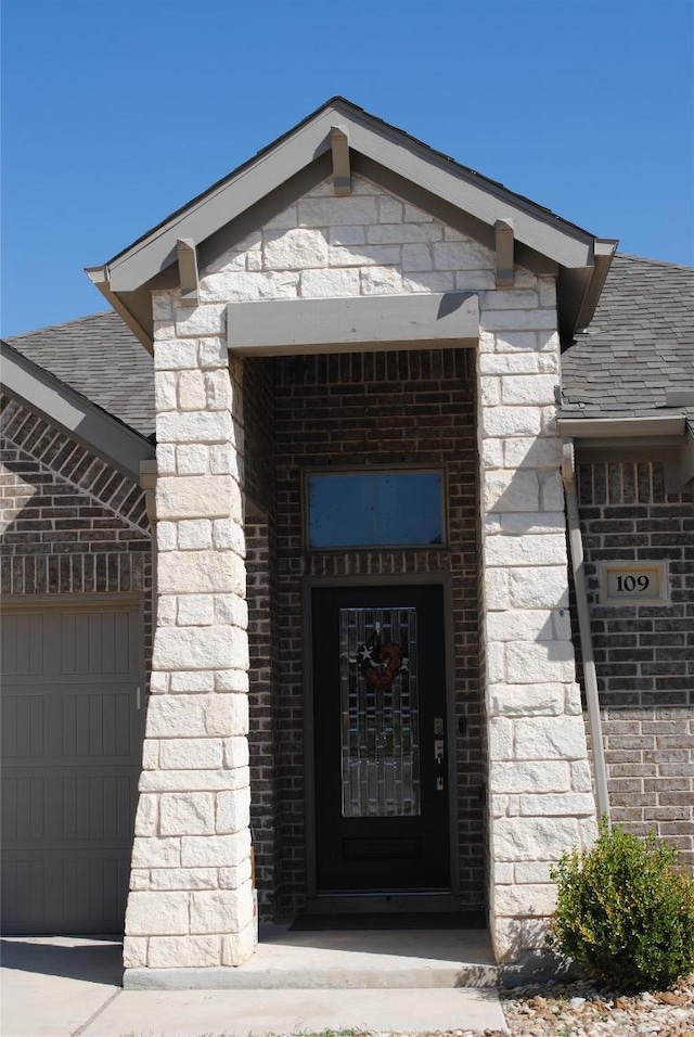 doorway to property featuring a shingled roof, brick siding, and an attached garage