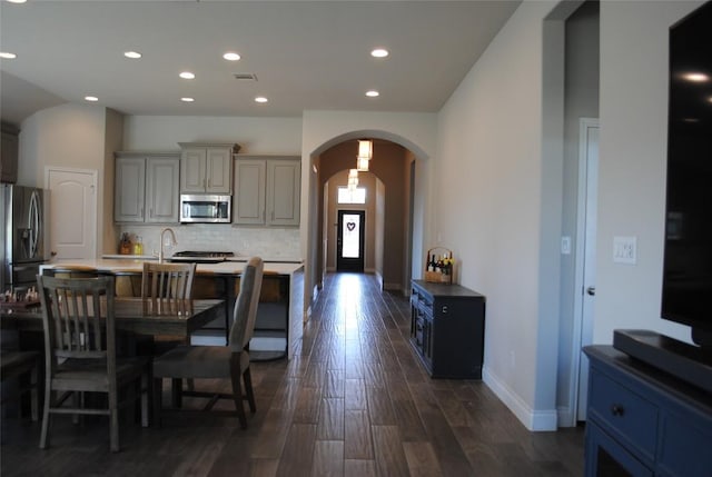 kitchen with arched walkways, dark wood-style flooring, a center island with sink, tasteful backsplash, and appliances with stainless steel finishes