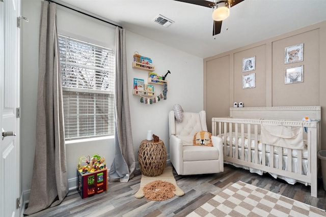 bedroom with a crib, wood finished floors, visible vents, and a ceiling fan