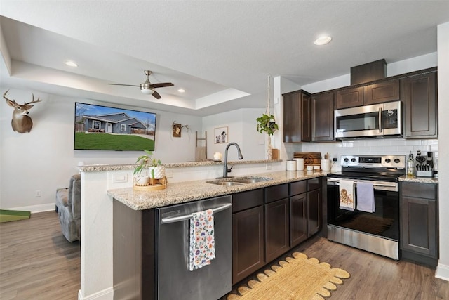 kitchen featuring a tray ceiling, a peninsula, a sink, dark brown cabinetry, and appliances with stainless steel finishes