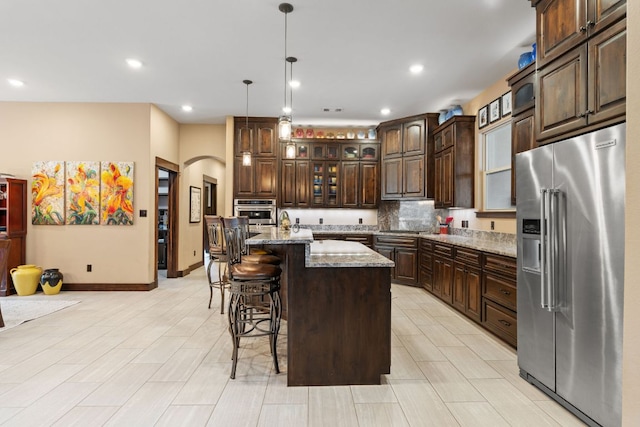 kitchen with a center island with sink, arched walkways, dark brown cabinetry, appliances with stainless steel finishes, and a kitchen breakfast bar