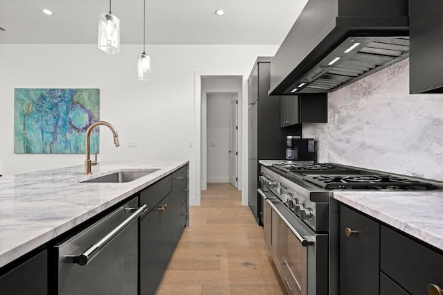 kitchen with dark cabinetry, a sink, stainless steel appliances, wall chimney exhaust hood, and light wood-type flooring