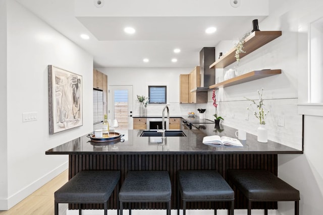 kitchen featuring a peninsula, light wood-style flooring, a sink, wall chimney range hood, and tasteful backsplash