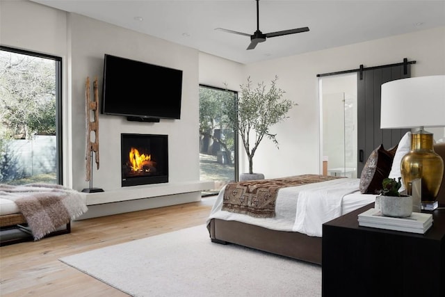 bedroom featuring a ceiling fan, wood finished floors, a warm lit fireplace, a barn door, and connected bathroom