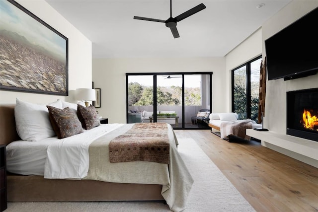 bedroom with a ceiling fan, access to outside, wood finished floors, and a warm lit fireplace