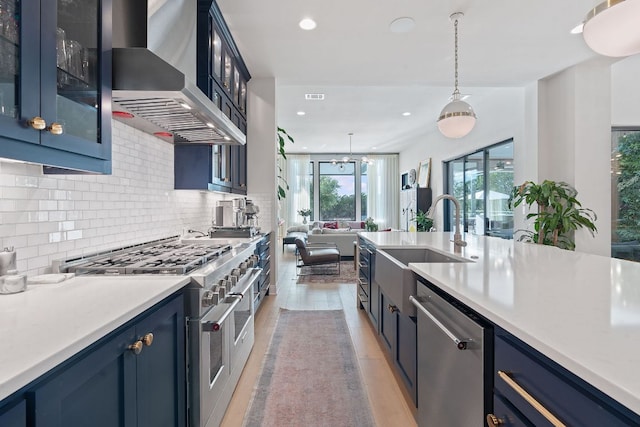 kitchen with blue cabinetry, a sink, backsplash, appliances with stainless steel finishes, and wall chimney range hood