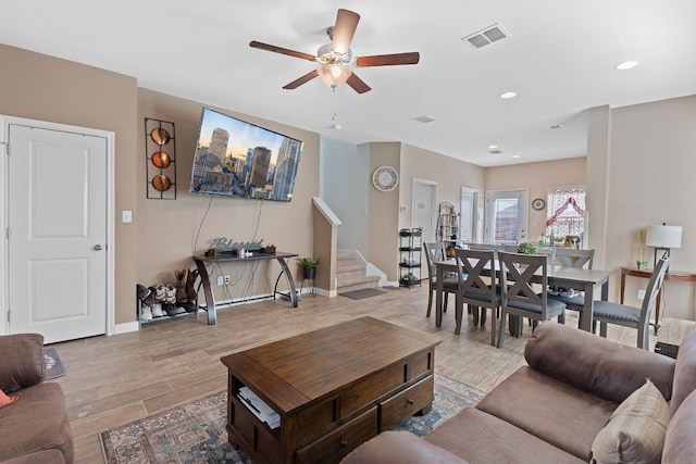 living room with visible vents, a ceiling fan, light wood finished floors, baseboards, and stairs