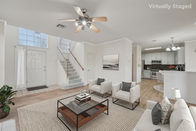 living room with visible vents, stairway, ornamental molding, ceiling fan with notable chandelier, and light wood-style floors