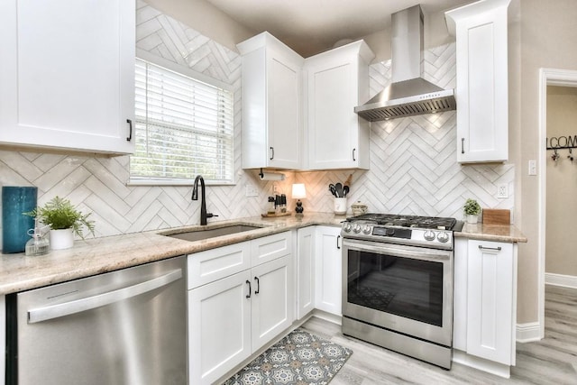 kitchen with a sink, wall chimney range hood, appliances with stainless steel finishes, white cabinets, and light wood finished floors