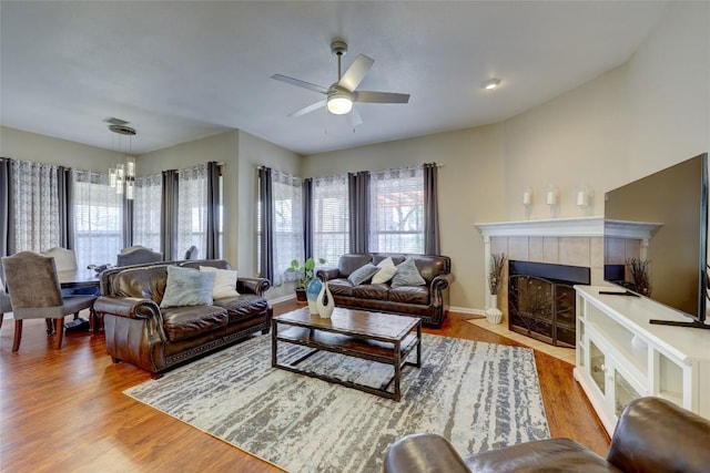 living room featuring baseboards, wood finished floors, a tiled fireplace, and ceiling fan with notable chandelier