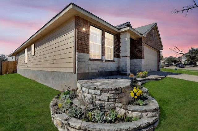 property exterior at dusk with driveway, stone siding, fence, a yard, and a garage