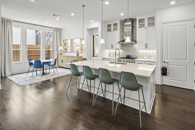 kitchen with a center island with sink, a sink, dark wood finished floors, wall chimney range hood, and decorative backsplash