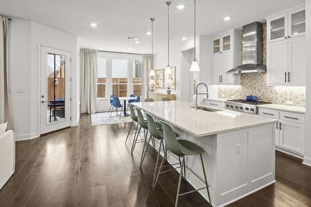 kitchen featuring tasteful backsplash, a sink, wall chimney range hood, a center island with sink, and dark wood-style flooring