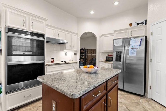 kitchen featuring arched walkways, stainless steel appliances, decorative backsplash, under cabinet range hood, and white cabinetry