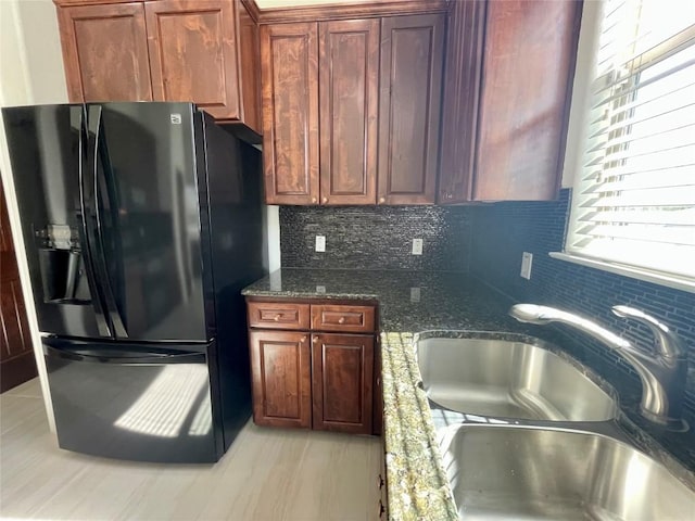 kitchen with black fridge with ice dispenser, backsplash, dark stone counters, and a sink