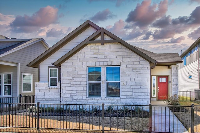 bungalow with stone siding, a fenced front yard, and a shingled roof