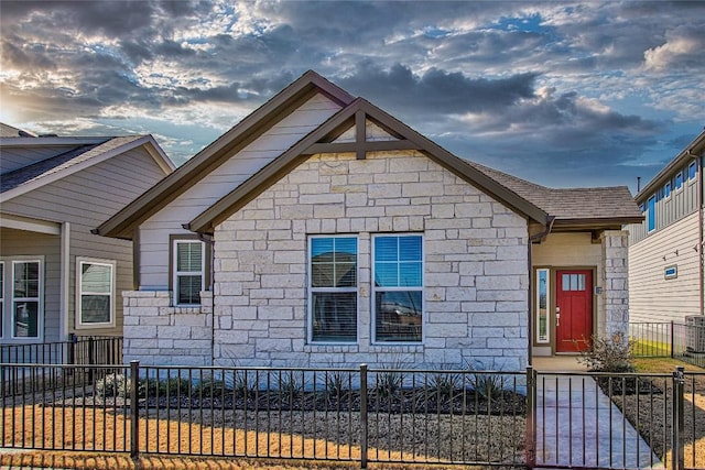 bungalow-style house featuring a fenced front yard, stone siding, and roof with shingles