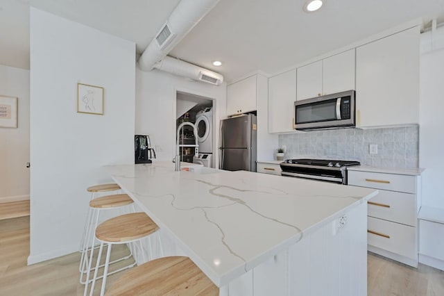 kitchen featuring light wood-style flooring, tasteful backsplash, appliances with stainless steel finishes, a breakfast bar area, and stacked washer / dryer