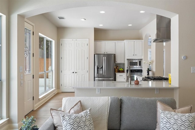 kitchen featuring light wood-type flooring, white cabinetry, appliances with stainless steel finishes, a peninsula, and wall chimney range hood