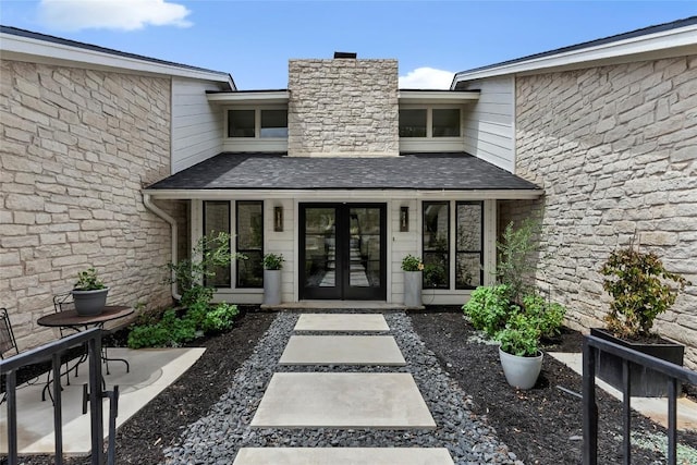 doorway to property featuring french doors, a chimney, stone siding, and roof with shingles