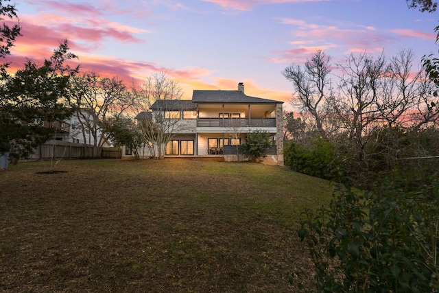 back of house at dusk featuring a yard, a chimney, a balcony, and fence