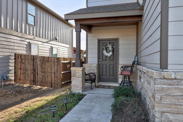 view of exterior entry with stone siding, a shingled roof, and fence