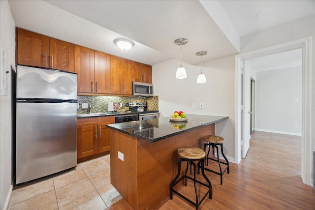 kitchen featuring stainless steel appliances, a peninsula, backsplash, a kitchen bar, and brown cabinetry