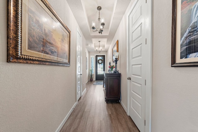 corridor featuring baseboards, wood finished floors, a textured wall, a notable chandelier, and a raised ceiling