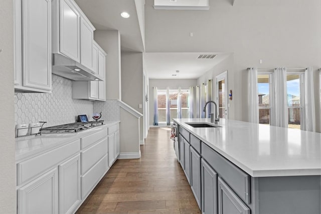 kitchen with wood finished floors, a kitchen island with sink, gray cabinets, a sink, and under cabinet range hood