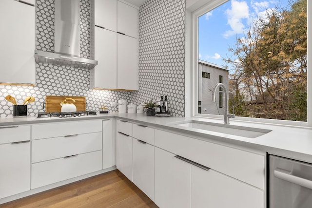 kitchen with light wood finished floors, a sink, wall chimney range hood, light countertops, and stainless steel dishwasher