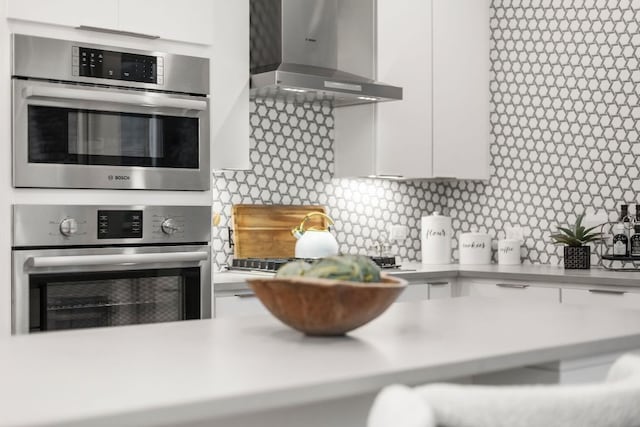 kitchen featuring double oven, white cabinetry, light countertops, and wall chimney range hood