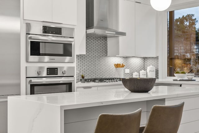 kitchen with light stone countertops, appliances with stainless steel finishes, wall chimney range hood, modern cabinets, and backsplash