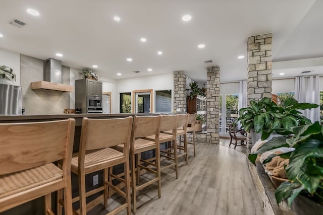 kitchen featuring recessed lighting, appliances with stainless steel finishes, wall chimney exhaust hood, light wood finished floors, and ornate columns